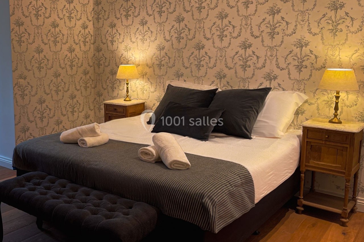 Lit double avec draps blancs, plaid gris, coussins noirs, tables de chevet en bois et lampes allumées dans une chambre cosy.