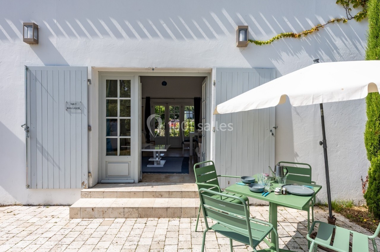 Terrasse ensoleillée avec table et chaises vertes, parasol blanc, et vue sur une porte-fenêtre ouverte d'une maison blanche.