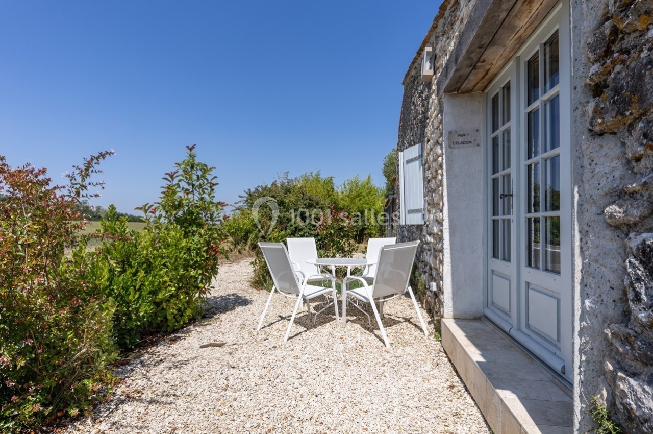 Table et chaises blanches sur une terrasse en gravier, entourées de verdure, devant une maison en pierre.