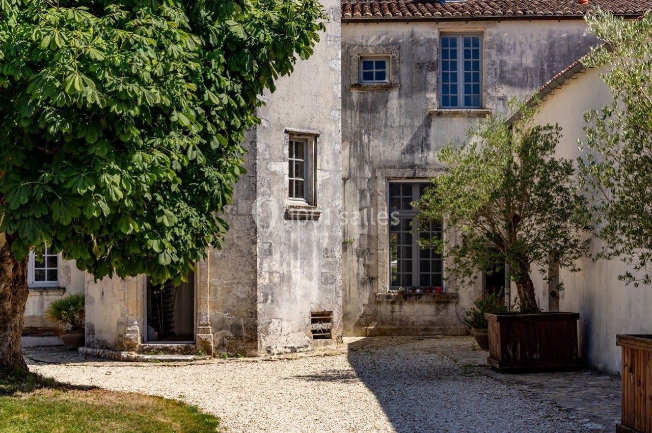Façade en pierre d'une maison ancienne avec fenêtres, arbres et cour pavée ensoleillée.