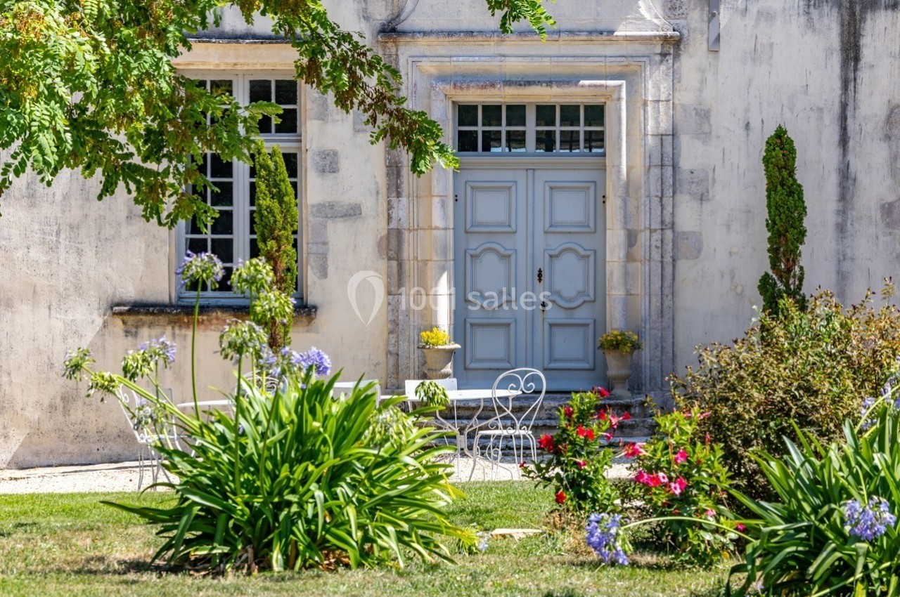 Façade d'une maison ancienne avec porte bleue, fenêtres à carreaux, jardin fleuri et mobilier de jardin blanc.