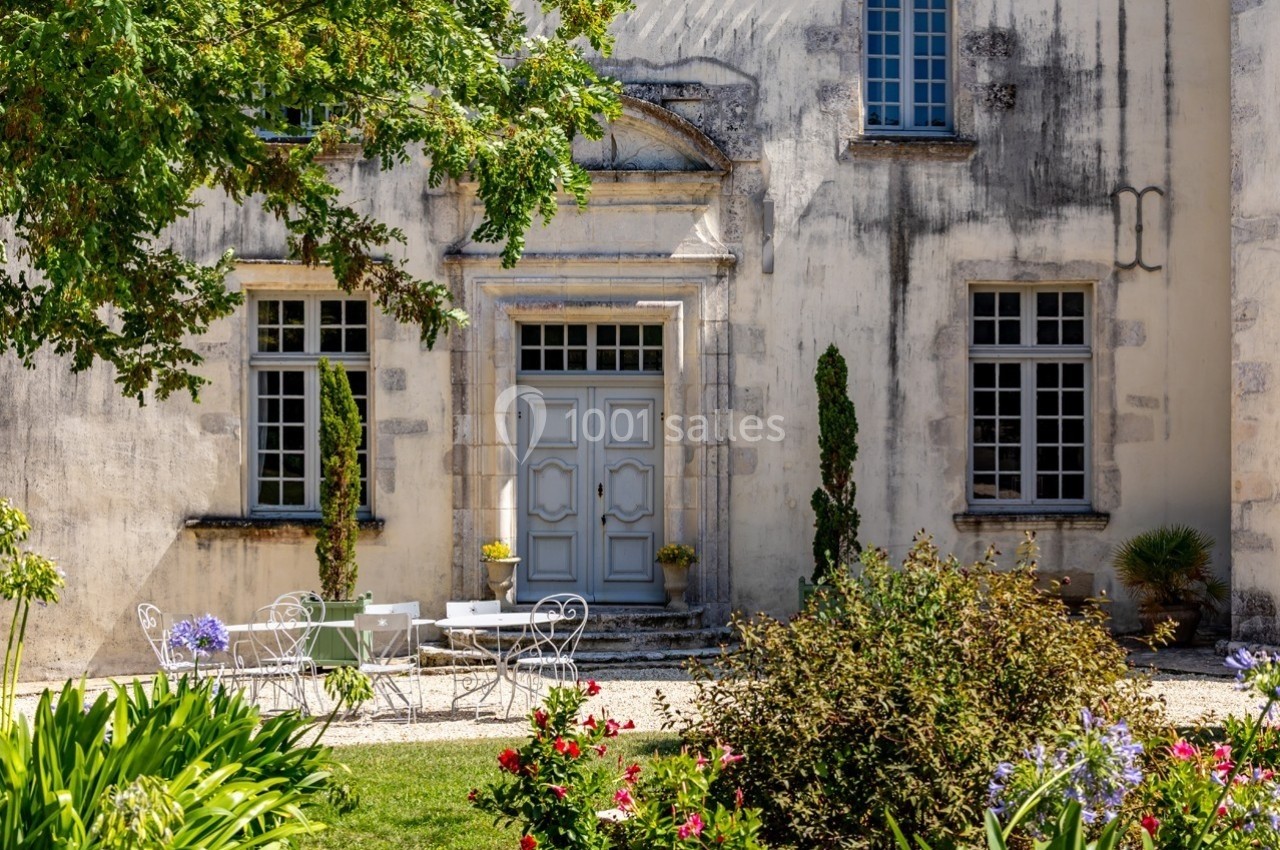 Façade d'un bâtiment ancien avec porte centrale, fenêtres symétriques, mobilier de jardin et végétation fleurie au premier…