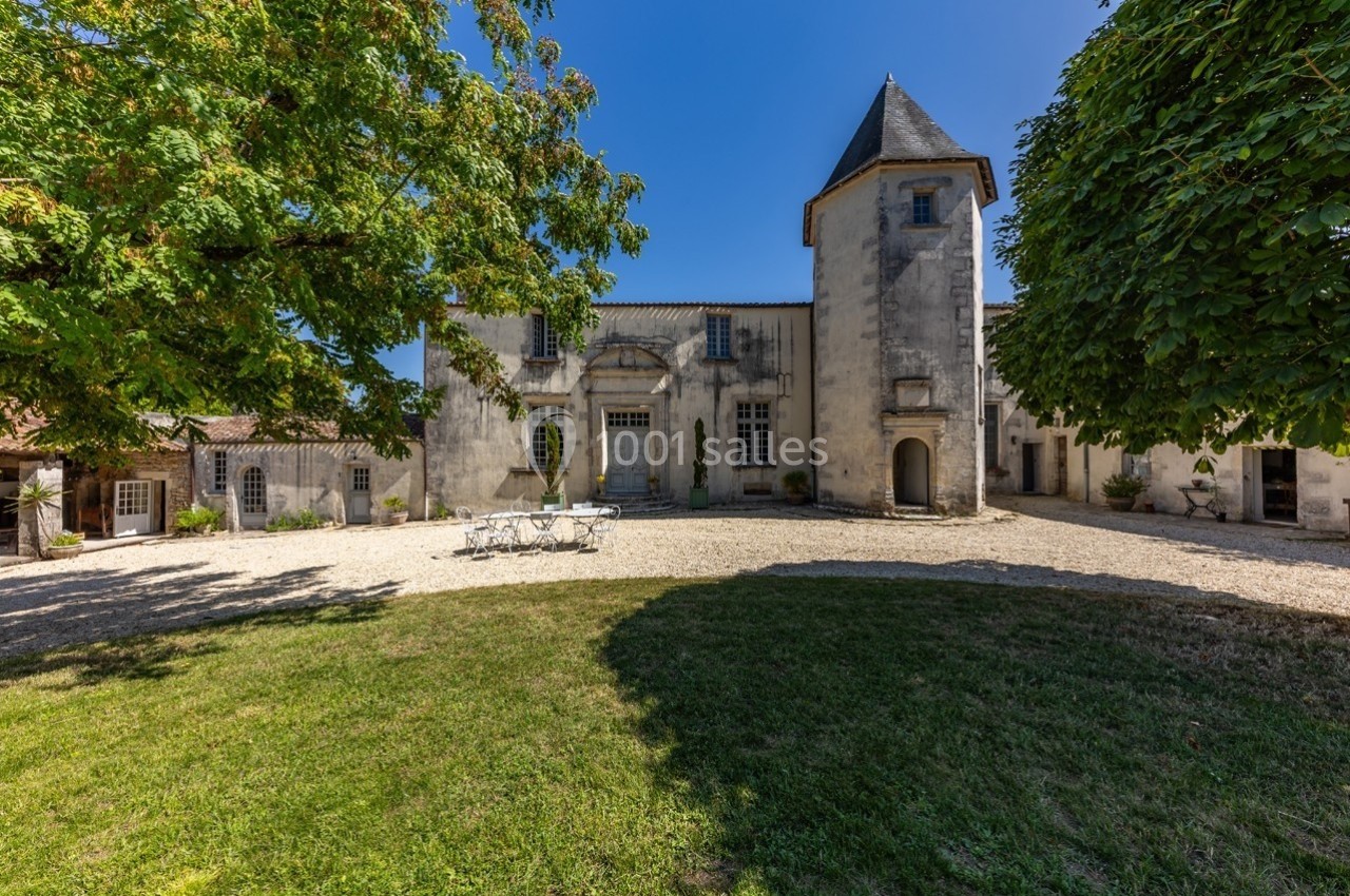 Façade d'un bâtiment ancien avec une tour, entouré de verdure et d'une cour en gravier sous un ciel bleu.