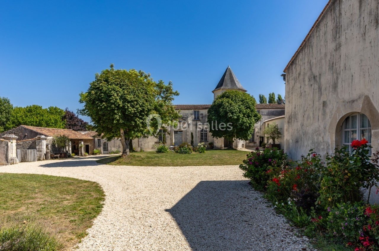 Cour d'un bâtiment ancien avec des murs en pierre, un chemin gravillonné, des arbres et des fleurs sous un ciel bleu.