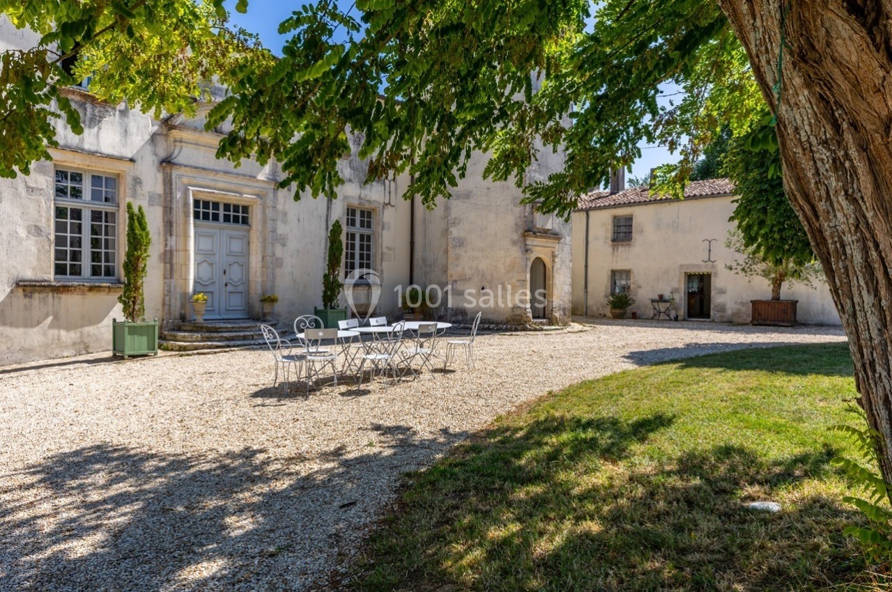 Cour d'un bâtiment ancien avec façade en pierre, mobilier de jardin blanc et arbres offrant de l'ombre.