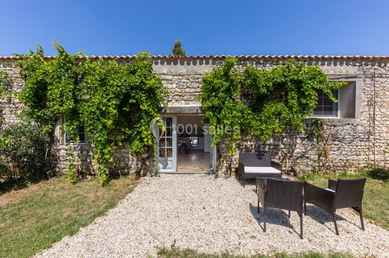 Façade en pierre d'une maison avec des plantes grimpantes, une porte ouverte et un salon de jardin sur gravier.