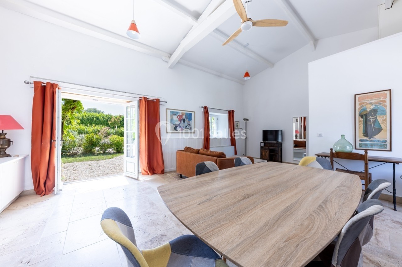 Salle à manger lumineuse avec table en bois, chaises colorées, canapé, et vue sur un jardin par une porte-fenêtre ouverte.