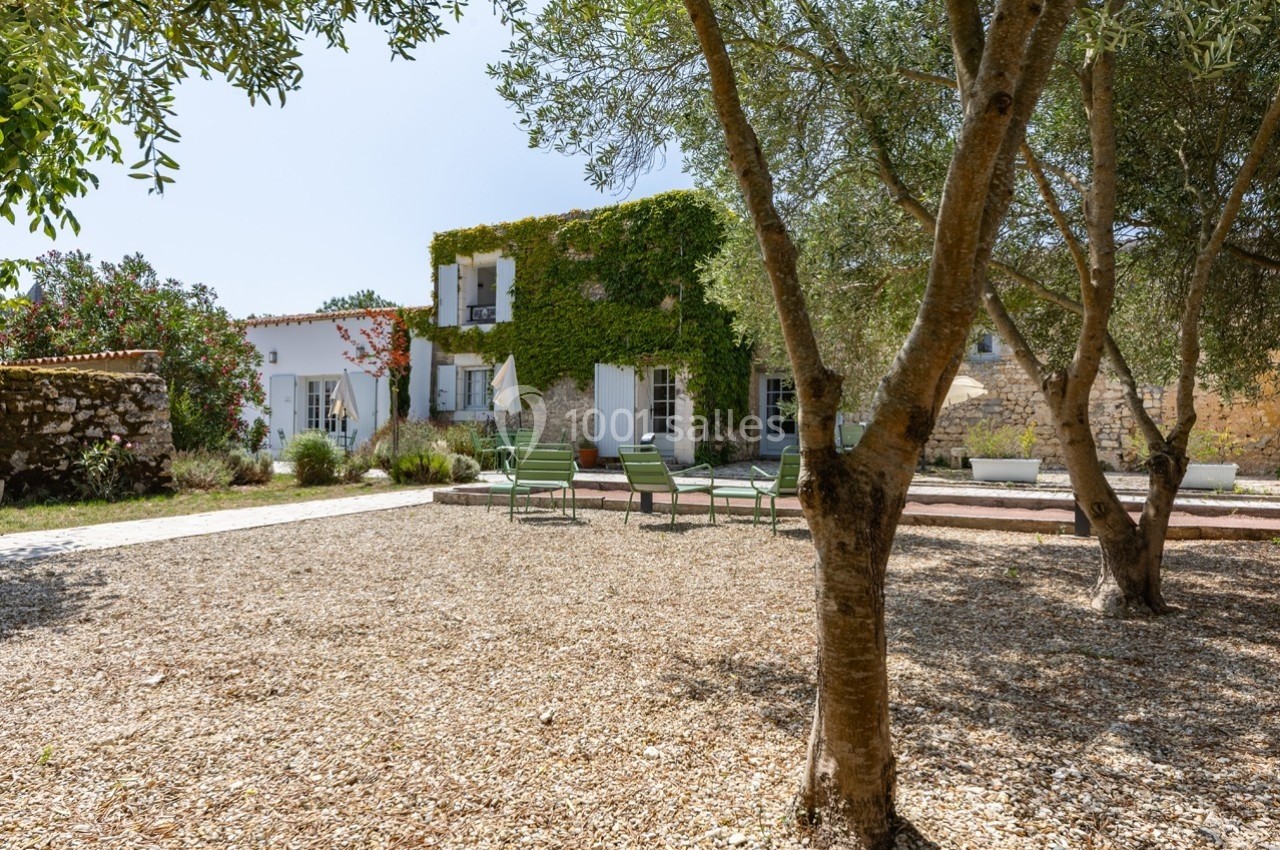 Maison en pierre recouverte de végétation, entourée d'arbres et d'une cour en gravier avec des chaises vertes.