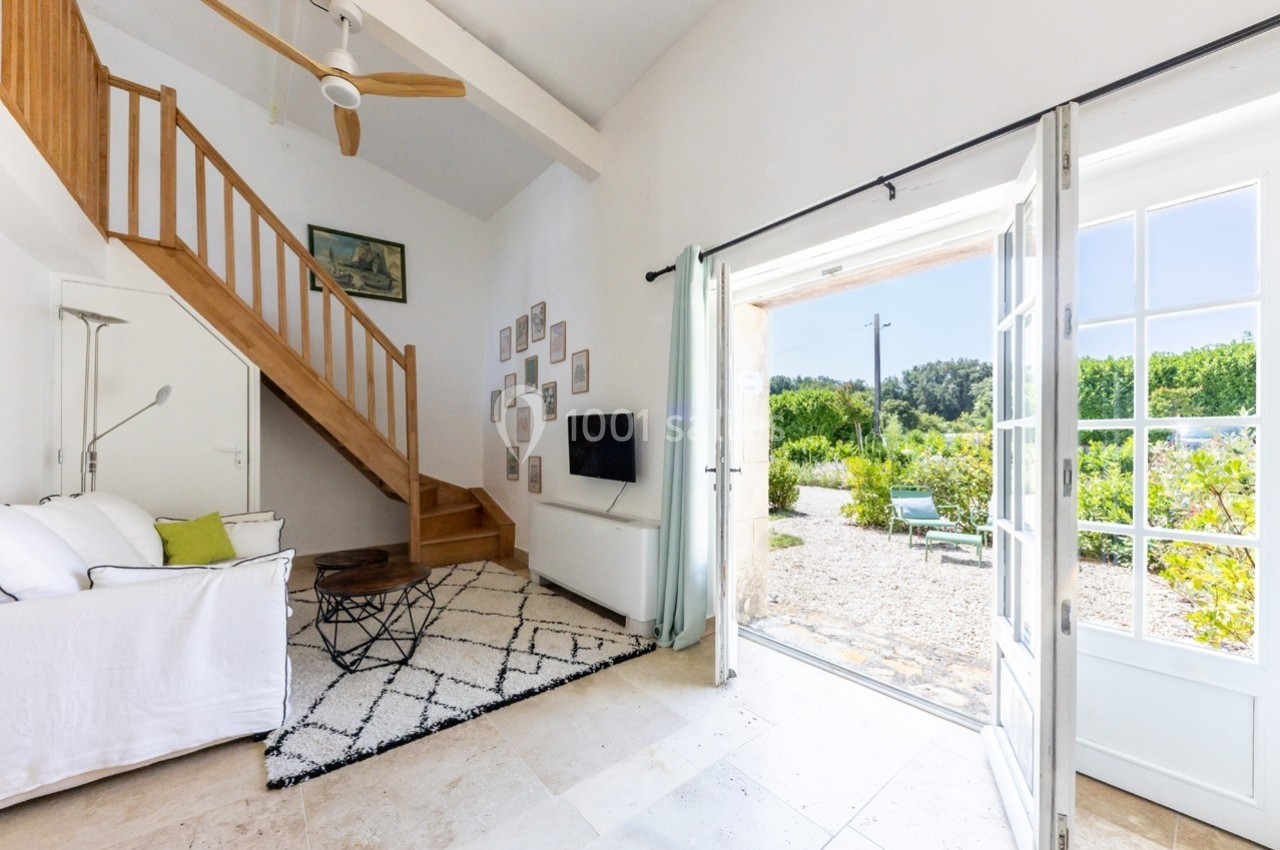 Salon lumineux avec canapé blanc, tapis géométrique, escalier en bois et porte-fenêtre ouverte sur un jardin.