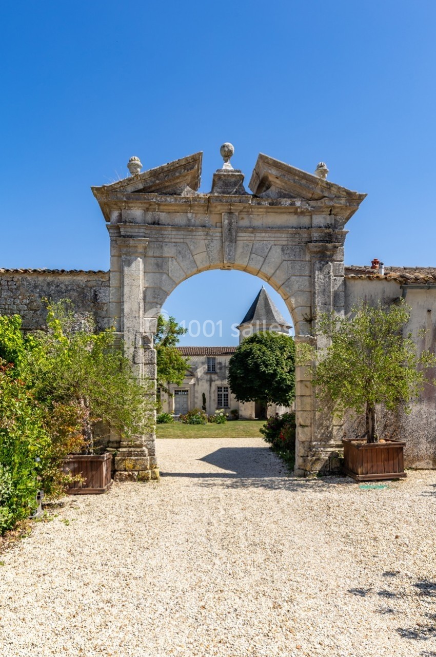 Porte en pierre ancienne avec une arche, donnant sur une cour pavée et un bâtiment avec un clocher sous un ciel bleu.