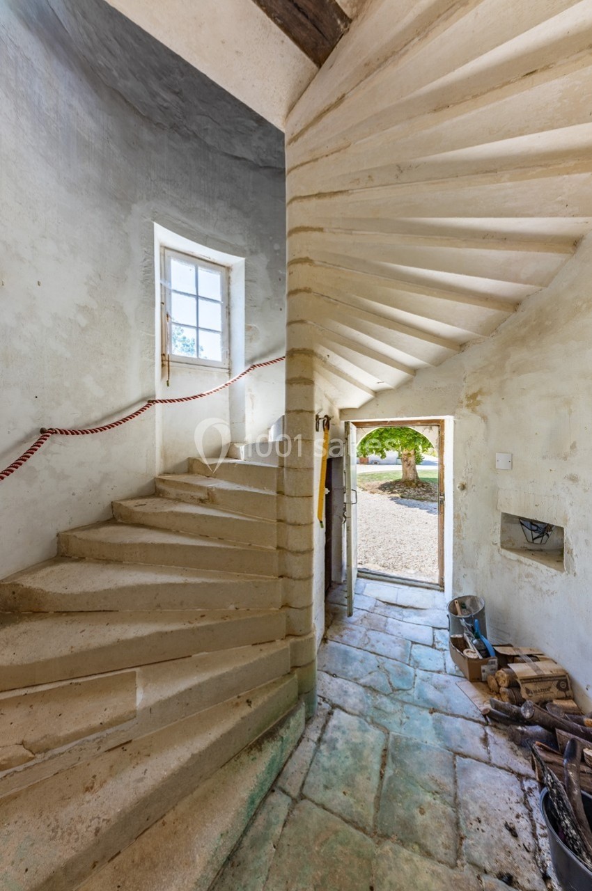 Escalier en pierre en colimaçon dans un intérieur rustique, avec une porte ouverte donnant sur une cour ensoleillée.