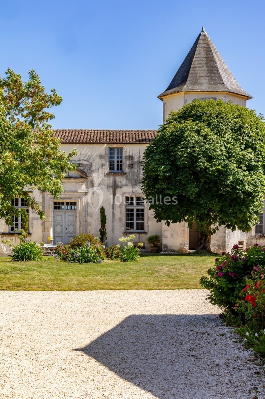 Façade d'une maison ancienne avec une tour, entourée d'arbres, d'un jardin et d'une allée en gravier.