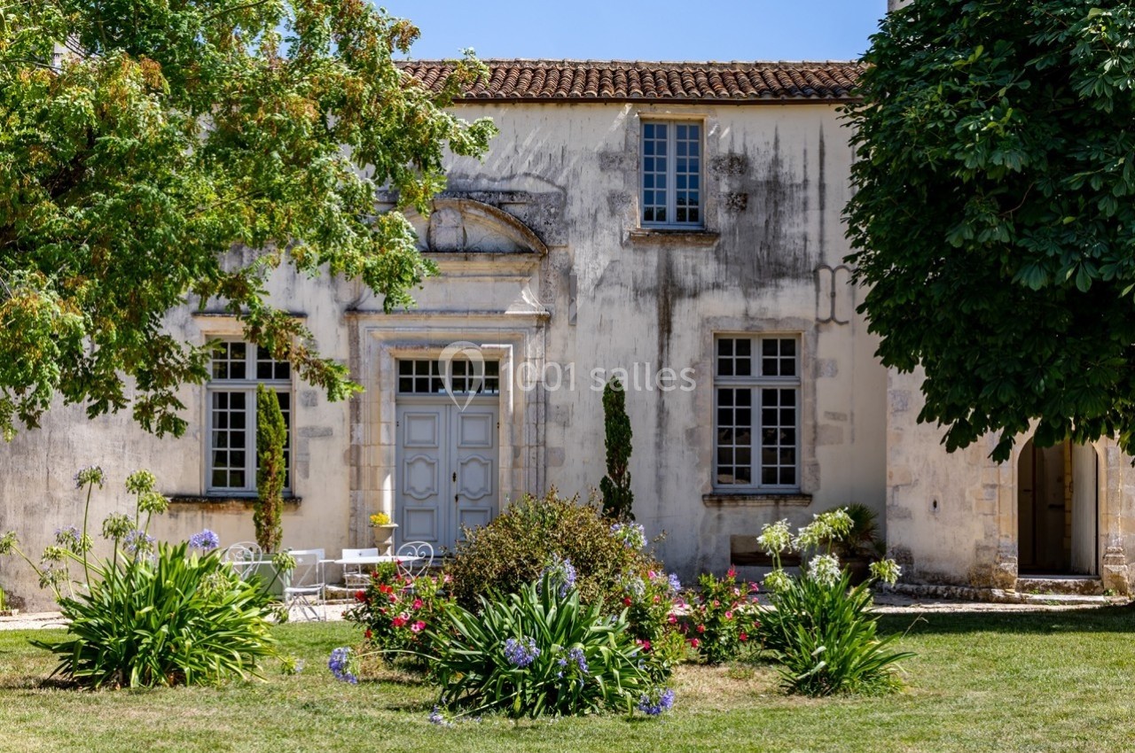 Façade d'une maison ancienne avec un jardin fleuri et des arbres sous un ciel bleu.