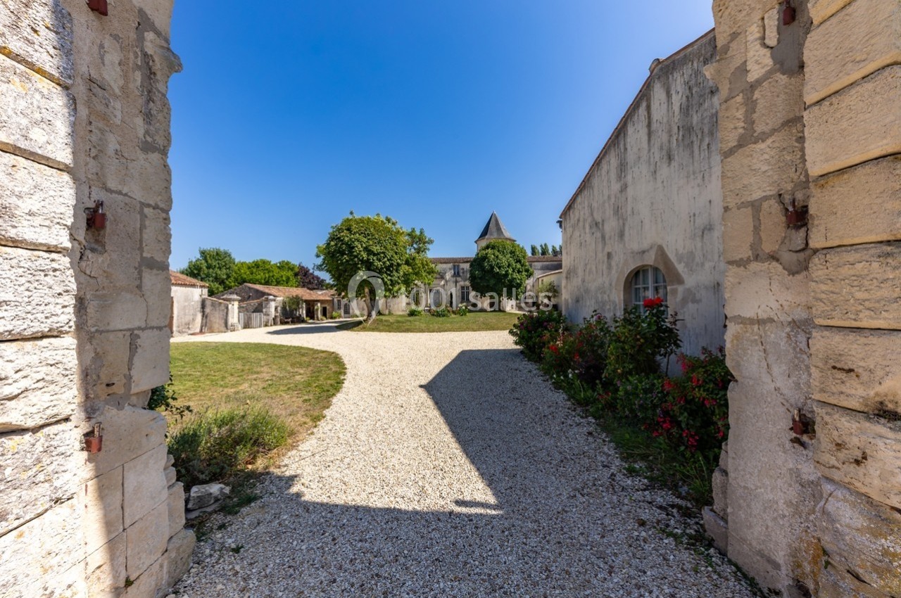 Cour intérieure d'un domaine ancien avec allée gravillonnée, bâtiments en pierre et végétation sous un ciel bleu.