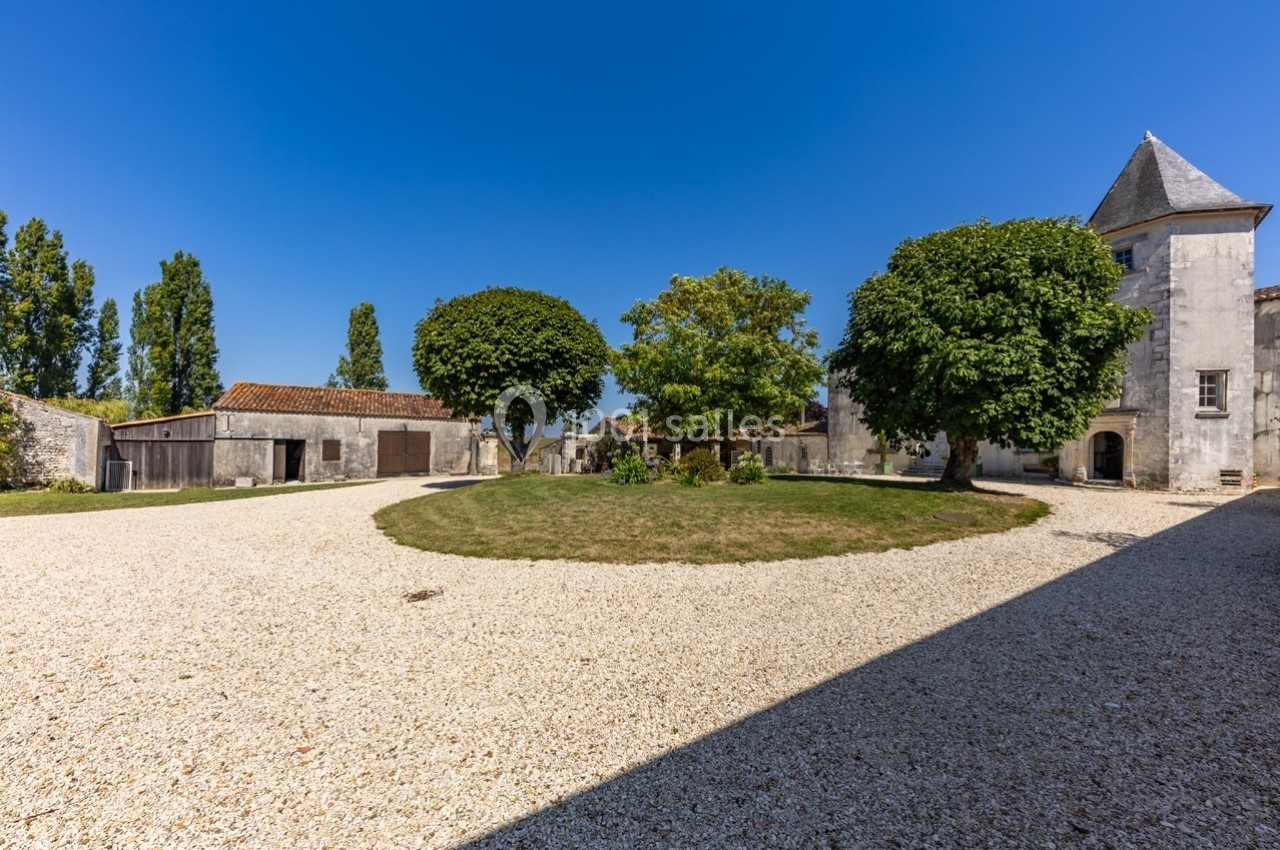 Cour d'une propriété avec gravier, pelouse centrale, arbres, bâtiments anciens en pierre et ciel bleu dégagé.
