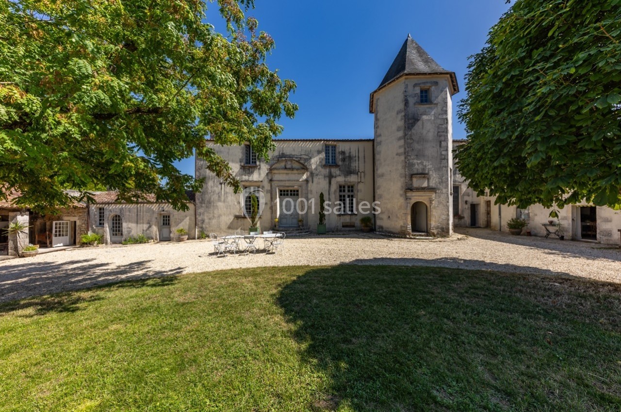 Façade d'un bâtiment ancien avec une tour, une cour gravillonnée et des arbres sous un ciel bleu.