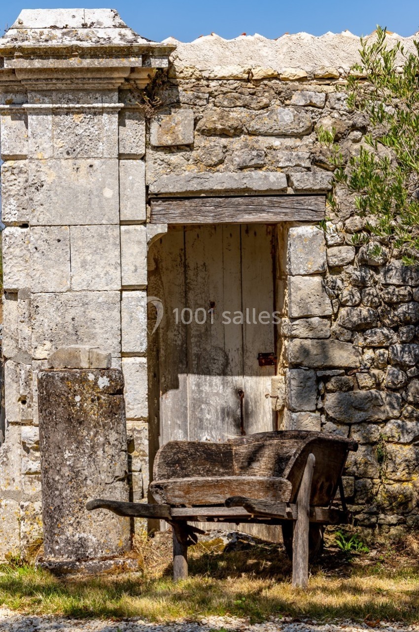 Vieille brouette en bois devant un mur en pierre et une porte en bois usée, sous un ciel dégagé.