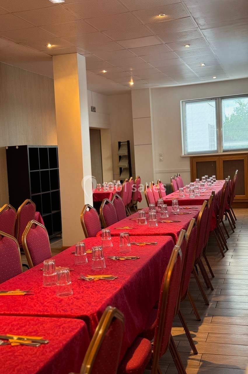 Salle aménagée avec des tables longues recouvertes de nappes rouges, alignées avec des chaises rouges et des couverts…