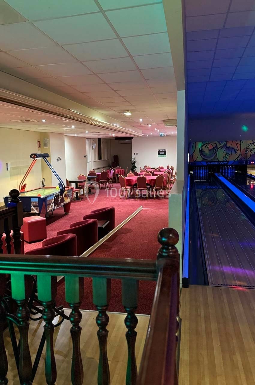 Salle de bowling avec une piste éclairée à droite et un espace de détente avec tables et fauteuils rouges à gauche.