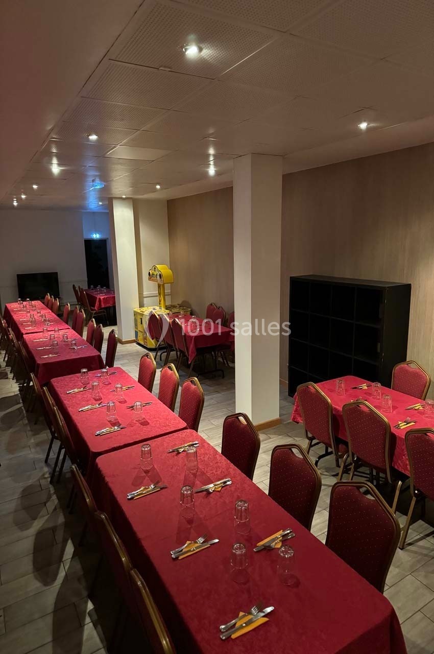 Salle de réception avec tables alignées, nappes rouges, chaises rouges et une machine de jeu au fond.