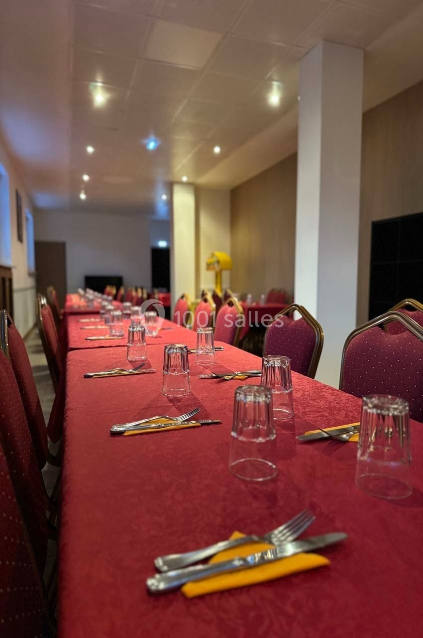 Salle avec de longues tables dressées de nappes rouges, couverts, verres et serviettes jaunes, prête pour un repas.