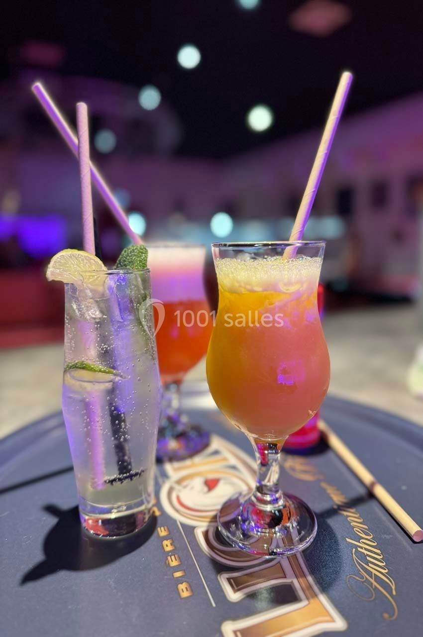 Trois cocktails colorés avec pailles en papier, servis sur un plateau dans un bar à l'ambiance lumineuse tamisée.