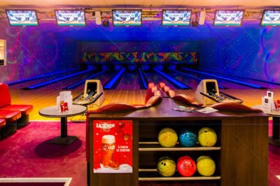 Boule de bowling jaune posée sur une piste en bois, avec des quilles alignées au fond et des lumières colorées.
