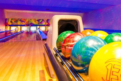 Boule de bowling jaune posée sur une piste en bois, avec des quilles alignées au fond et des lumières colorées.