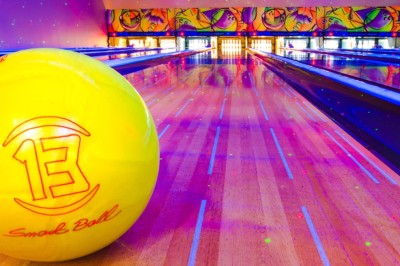 Boule de bowling jaune posée sur une piste en bois, avec des quilles alignées au fond et des lumières colorées.