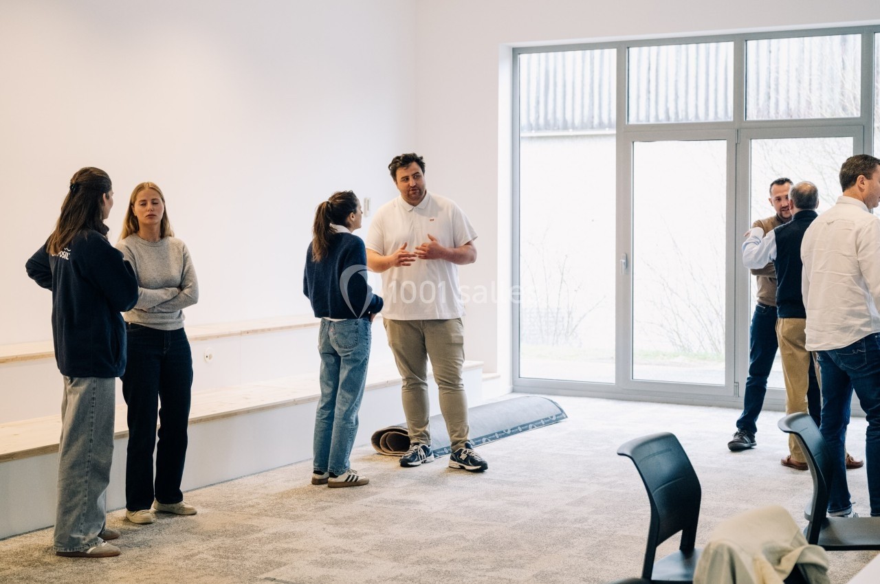 Des personnes discutent en petits groupes dans une salle lumineuse avec de grandes fenêtres et un sol recouvert de moquette.