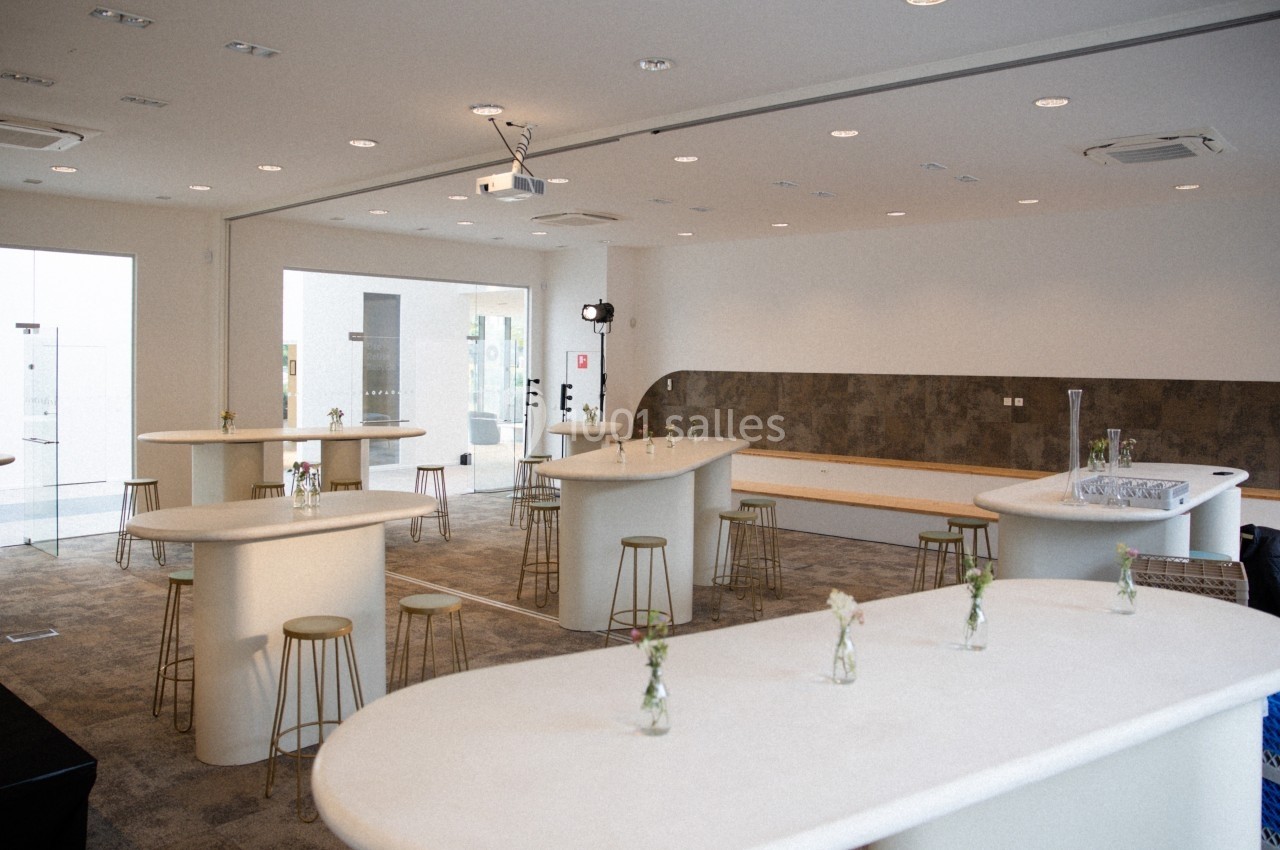 Salle lumineuse avec tables hautes blanches, tabourets, petites décorations florales et équipement audiovisuel.