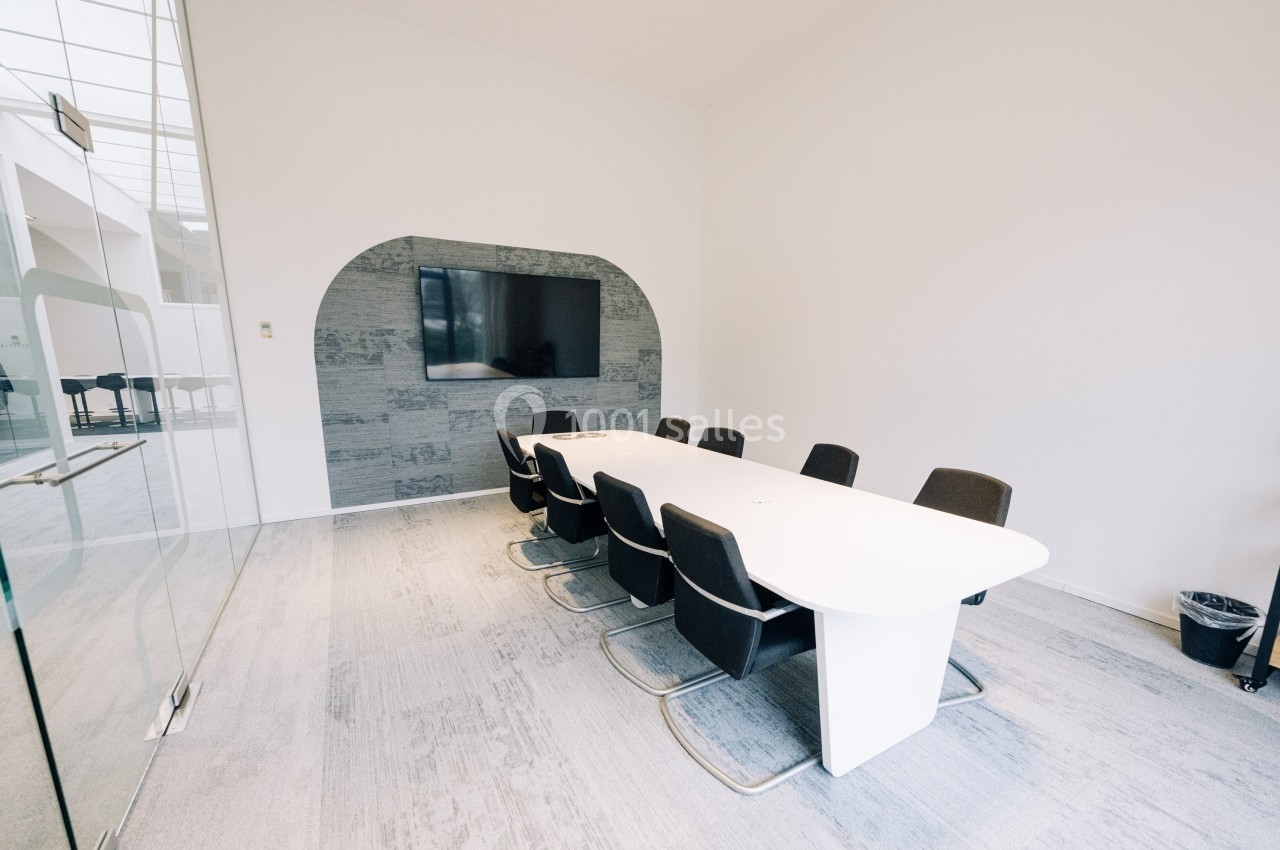 Salle de réunion moderne avec une grande table blanche, des chaises noires et un écran mural dans un espace lumineux.