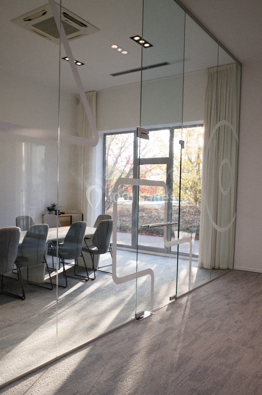 Salle de réunion moderne avec parois vitrées, table entourée de chaises grises et vue sur l'extérieur arboré.