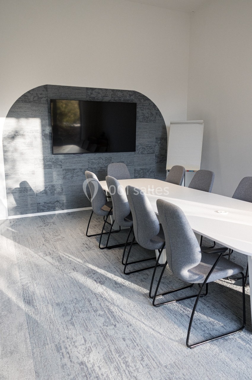 Salle de réunion lumineuse avec une table blanche, des chaises grises, un écran mural et un tableau de présentation.