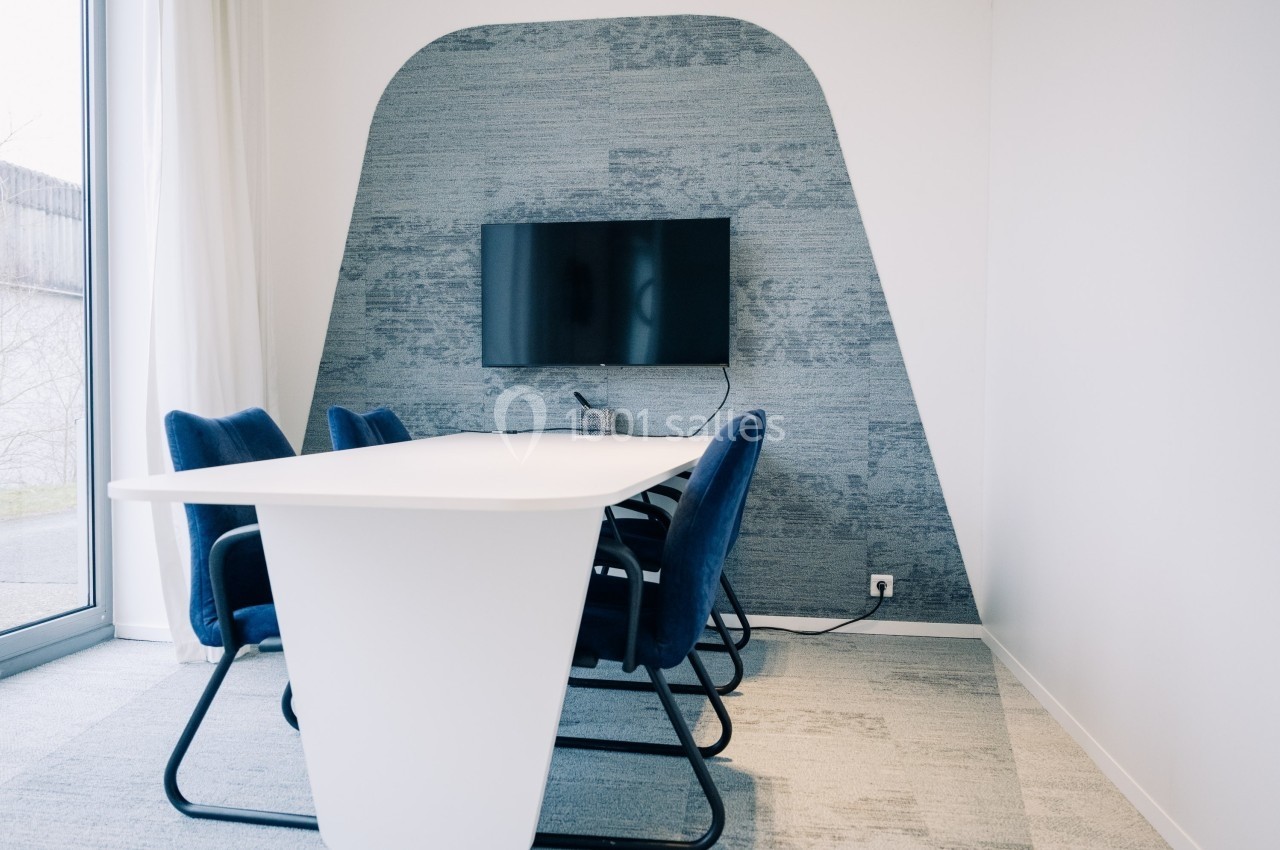Salle de réunion moderne avec table blanche, chaises bleues et écran mural sur fond de revêtement texturé.