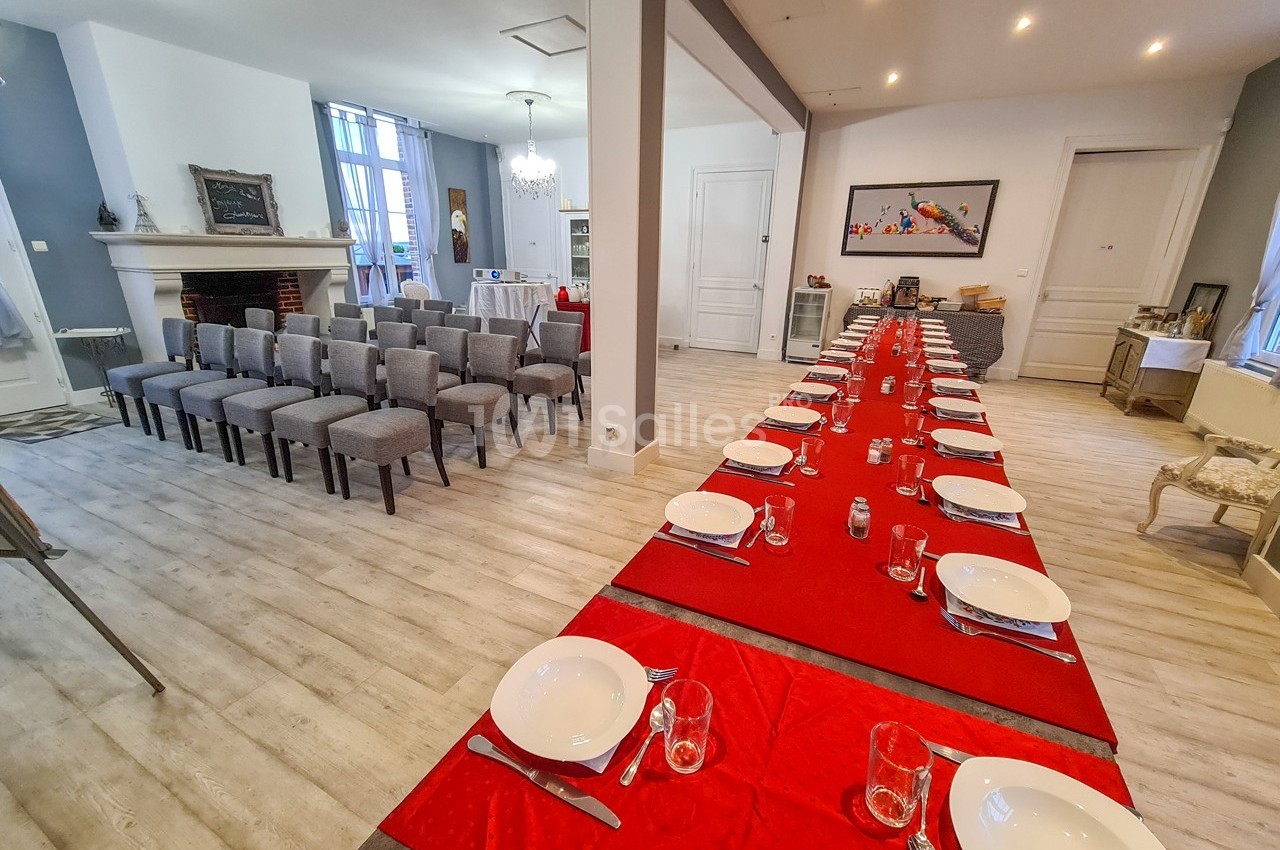 Salle lumineuse aménagée avec une longue table dressée et des rangées de chaises face à une cheminée.