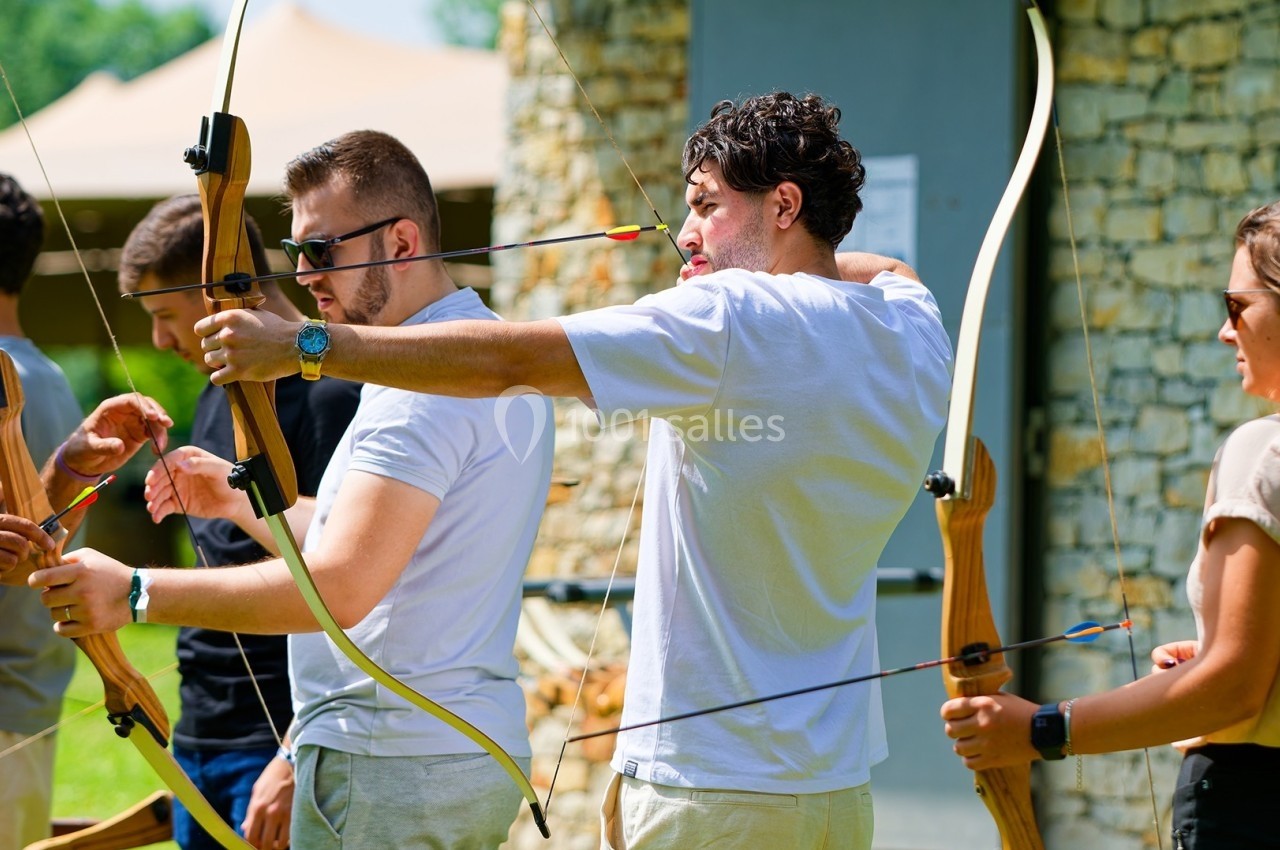 Des personnes apprennent le tir à l'arc en extérieur, encadrées par un instructeur près d'un mur en pierre.