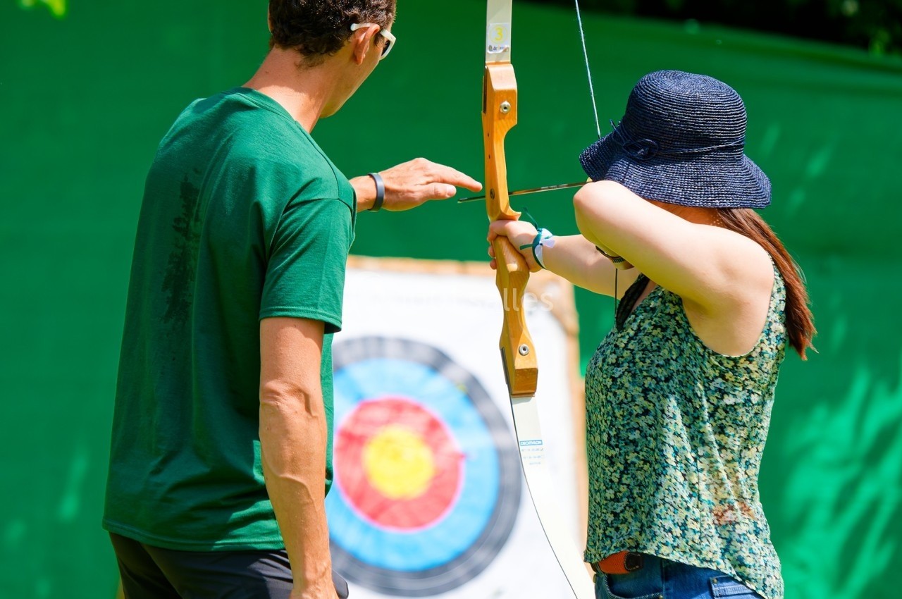 Une femme tire à l'arc sous la supervision d'un instructeur devant une cible en plein air.