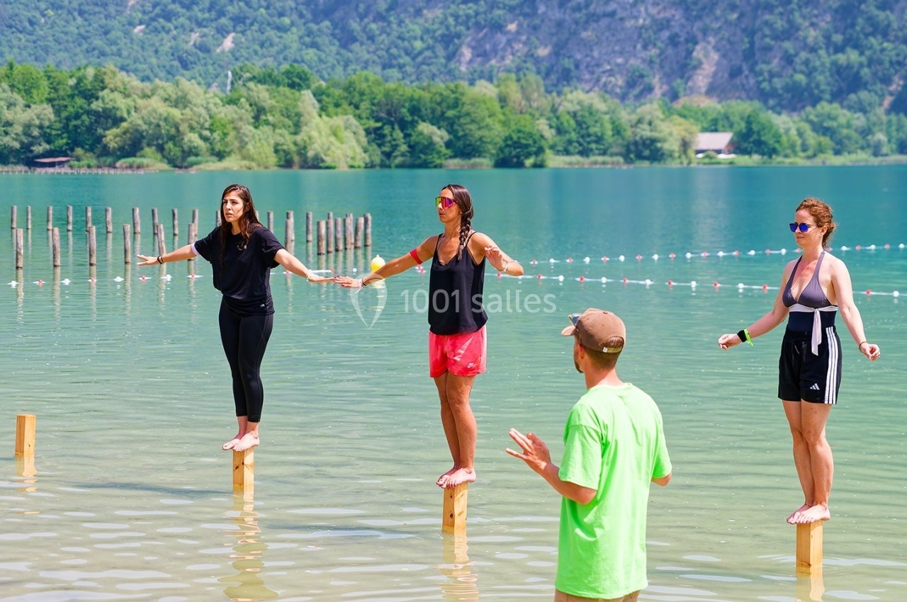 Trois femmes debout sur des poteaux dans un lac peu profond, encadrées par un homme en t-shirt vert, avec un paysage…