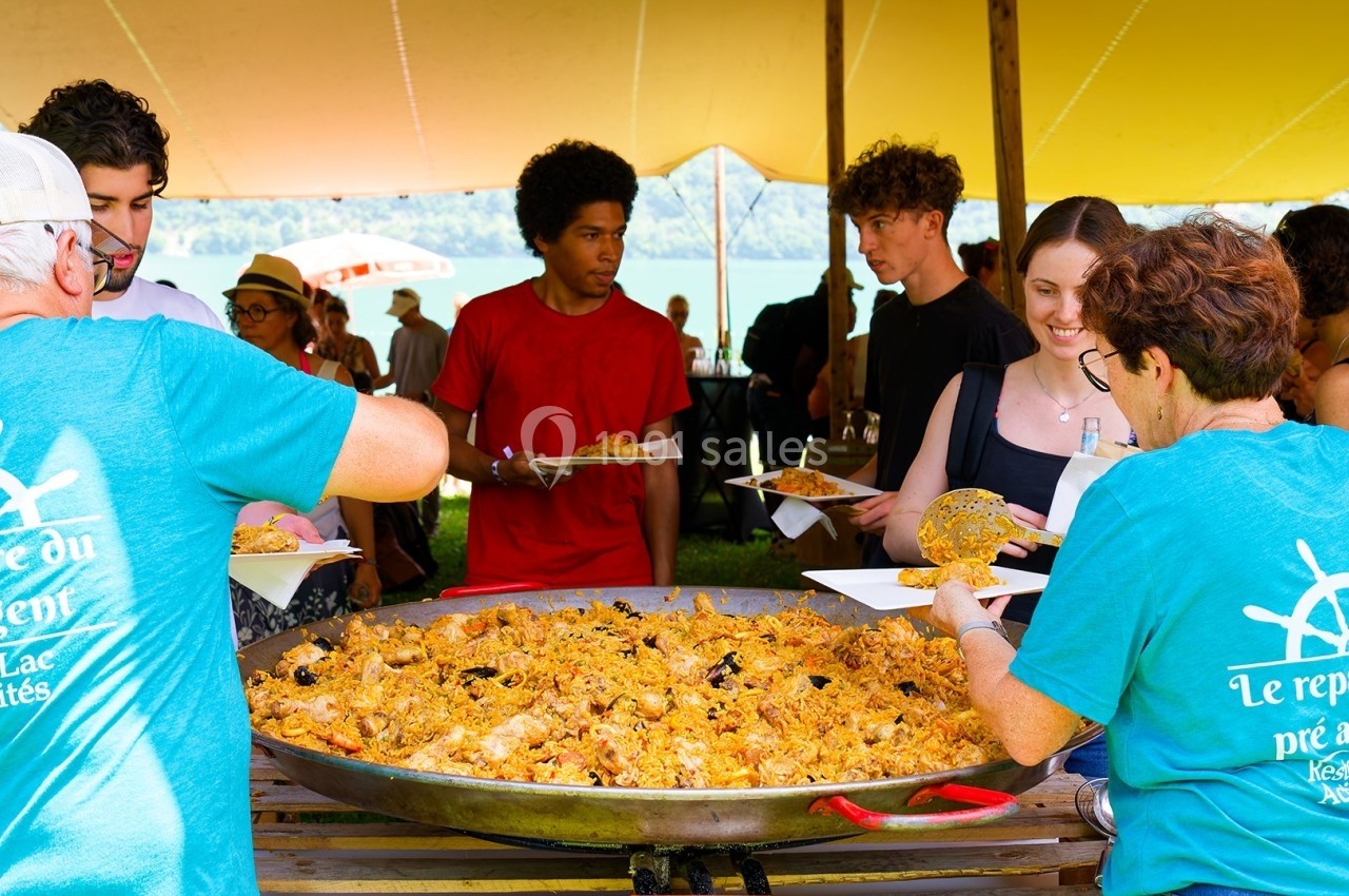 Des personnes se servent de la paella dans un grand plat lors d'un événement en plein air sous une tente.
