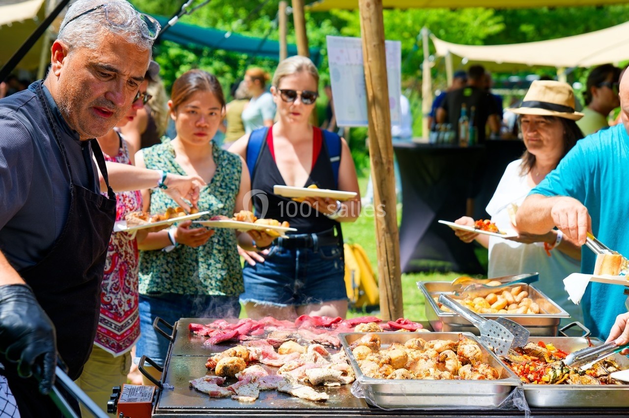 Un cuisinier prépare de la viande et des légumes sur une plancha lors d'un événement en plein air avec des participants.