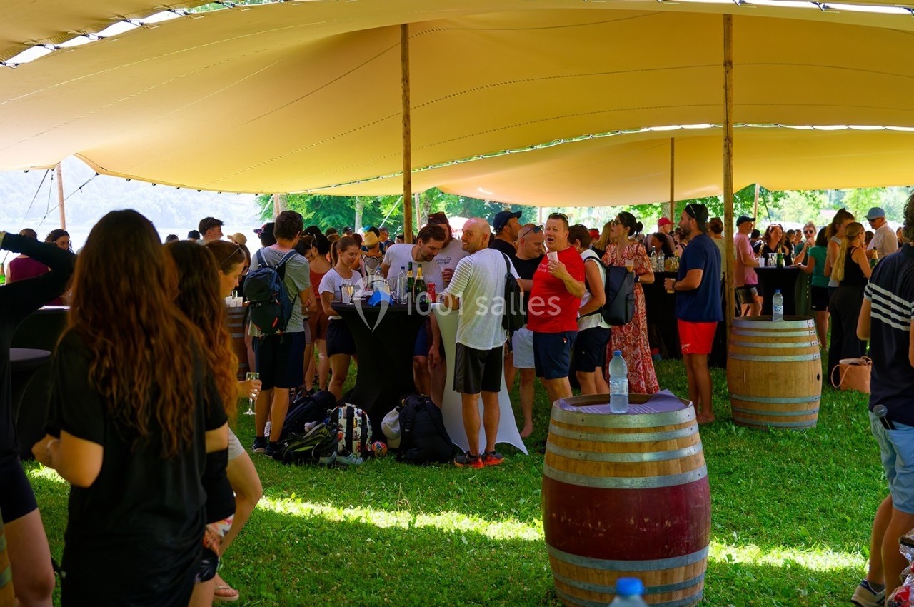 Groupe de personnes rassemblées sous une grande tente, discutant autour de tonneaux et de tables en plein air.