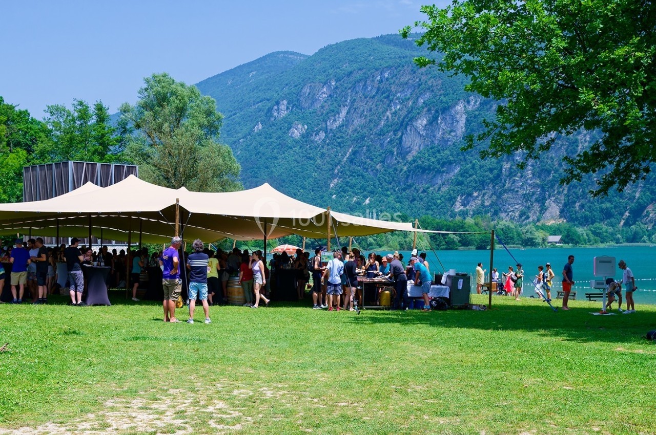 Groupe de personnes rassemblées sous une grande tente près d'un lac entouré de montagnes et de verdure.