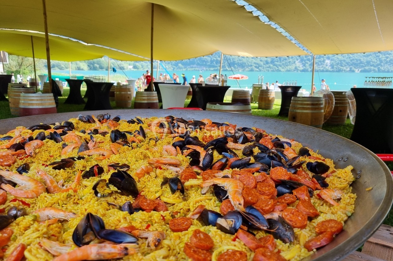 Grande poêle de paella garnie de crevettes, moules et chorizo, présentée sous une tente près d'un lac.