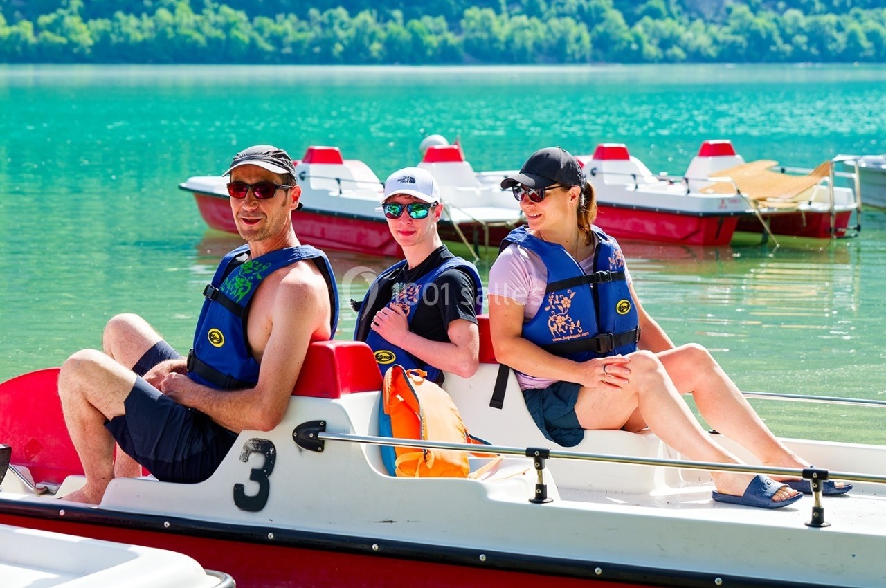 Trois personnes assises sur un pédalo au bord d'un lac turquoise, portant des gilets de sauvetage.