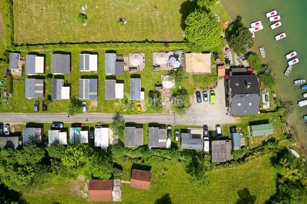 Vue aérienne d'un camping avec emplacements, bâtiments, voitures et accès direct à un lac avec des bateaux amarrés.