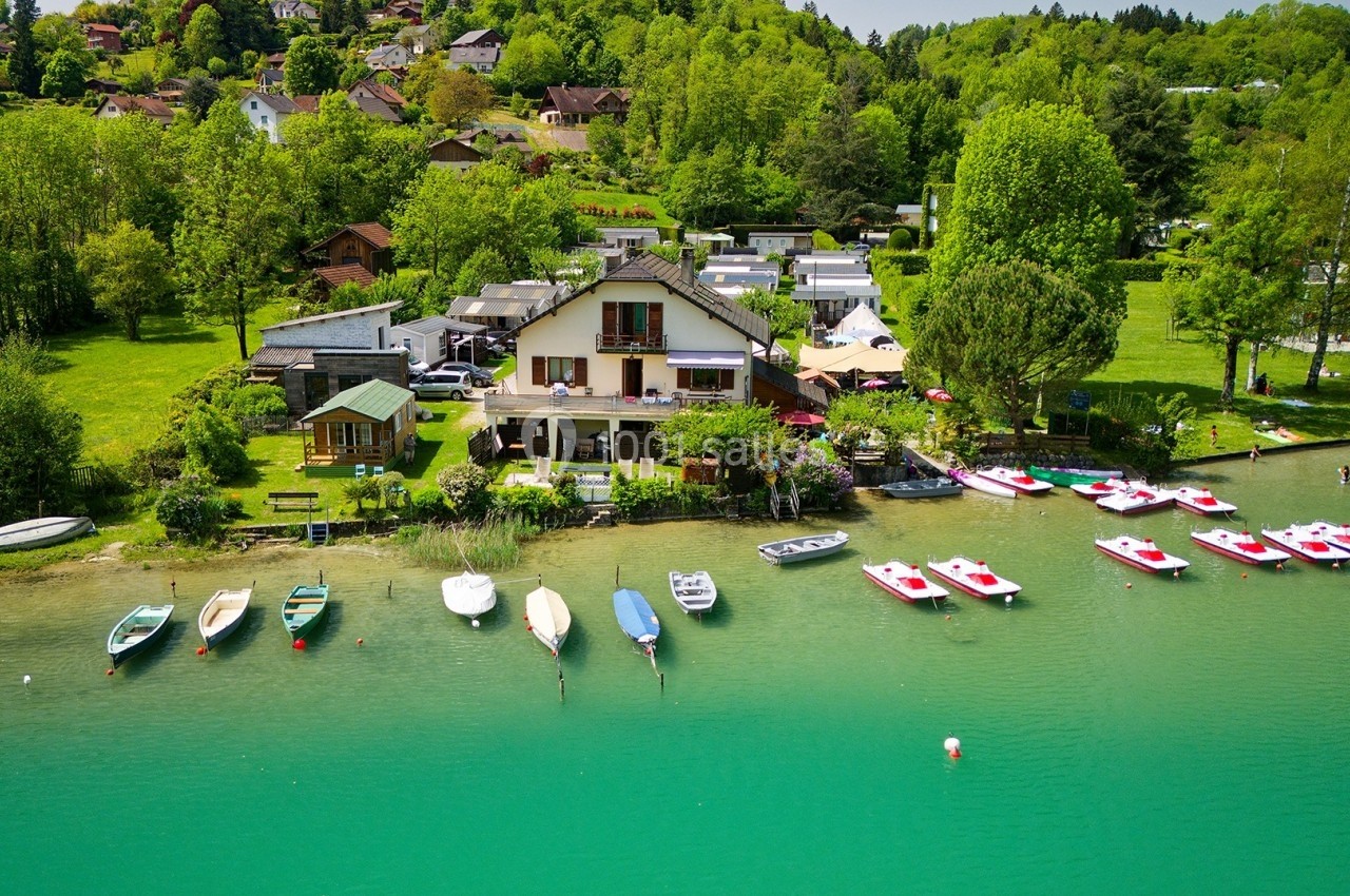 Vue d'une maison au bord d'un lac avec des bateaux amarrés et une végétation dense en arrière-plan.