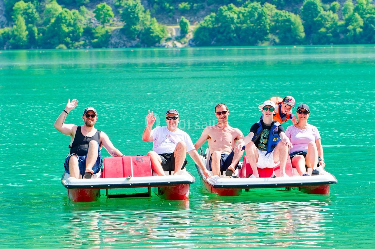 Un groupe de personnes souriantes sur deux pédalos rouges flottant sur un lac turquoise entouré de végétation.