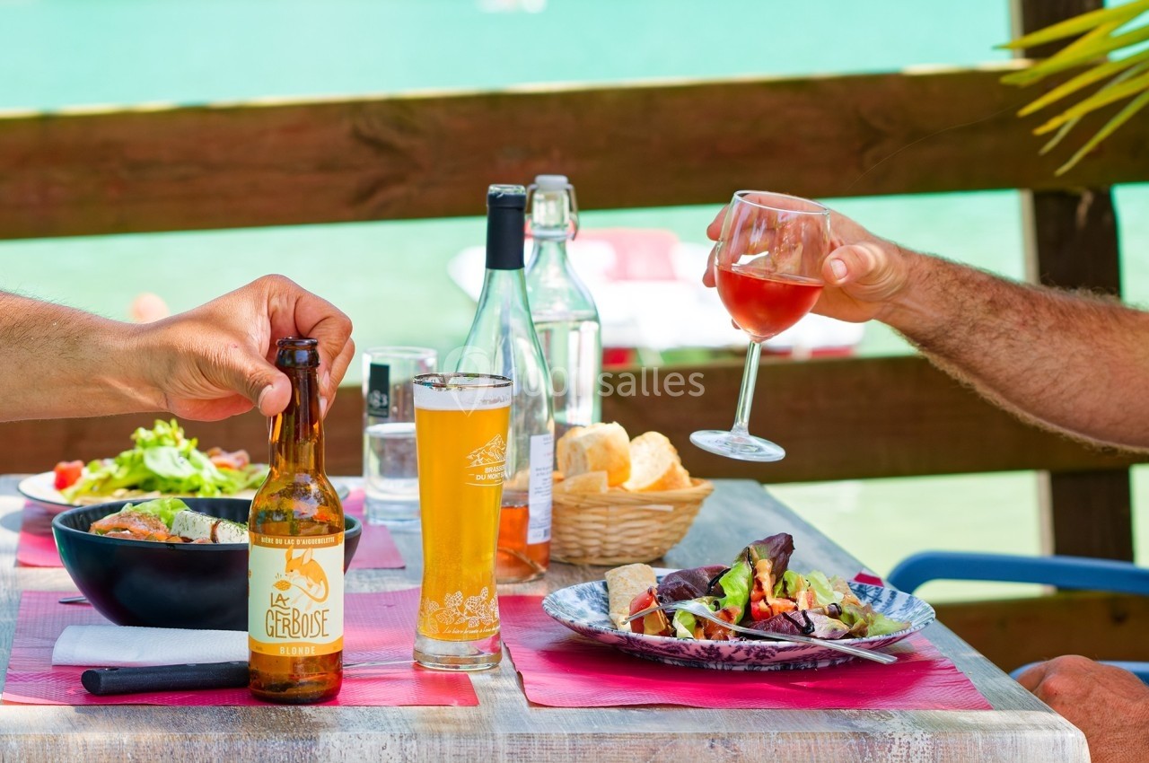 Repas en terrasse avec salades, pain, bière et vin, vue sur un lac en arrière-plan.