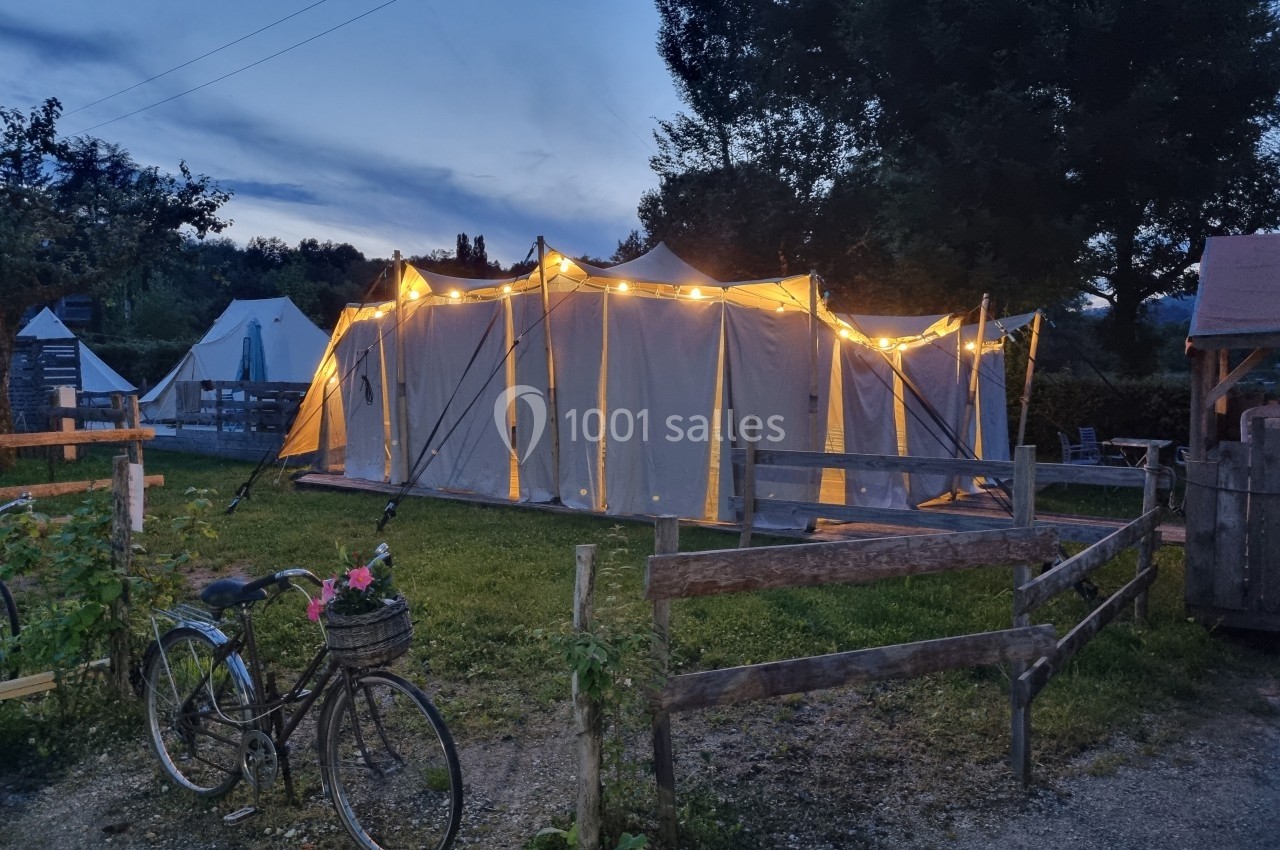 Tente éclairée au crépuscule dans un camping, avec une clôture en bois et un vélo garni d'un panier fleuri au premier plan.